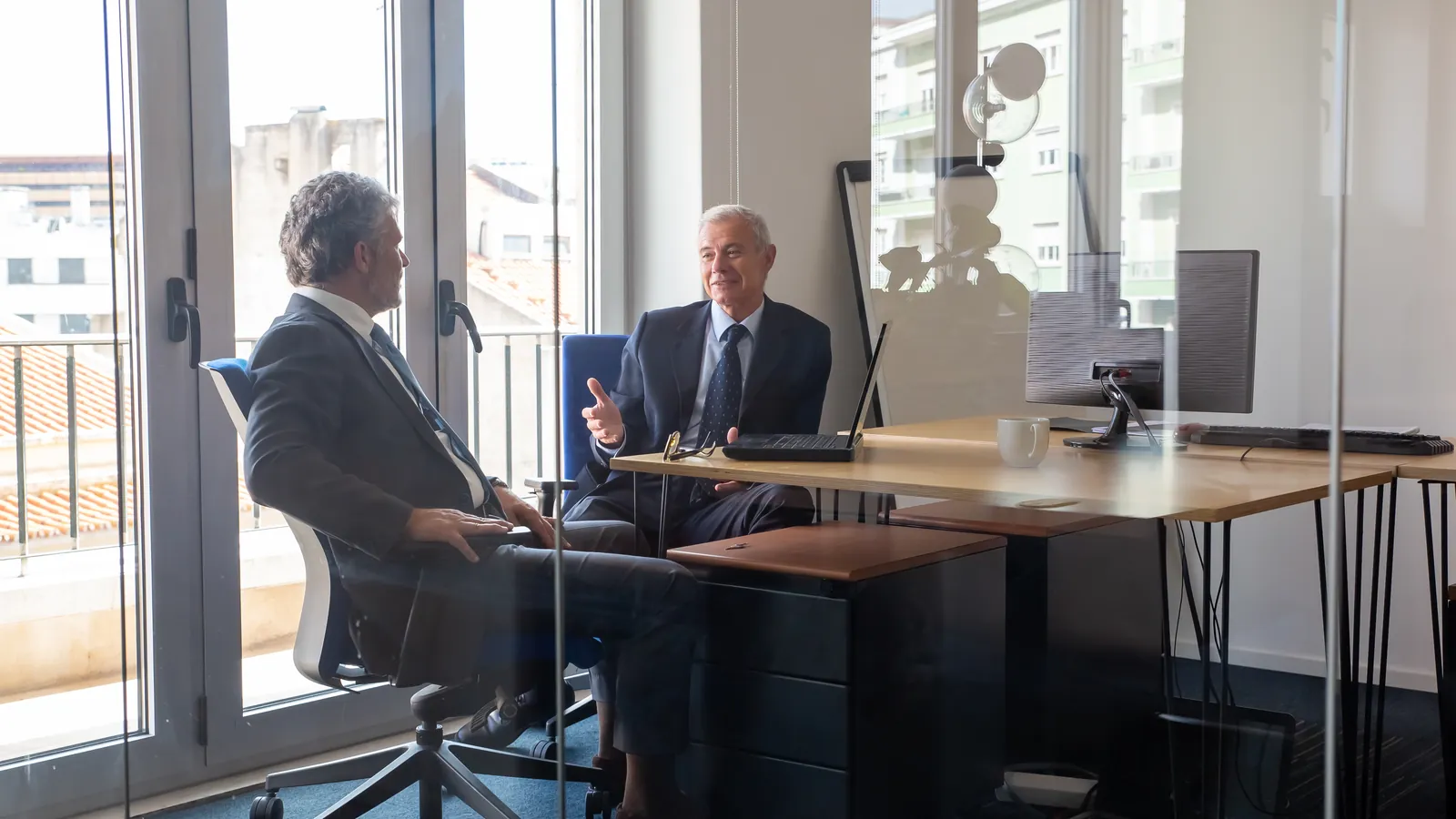 Dois sócios conversando numa sala de reunião com parede de vidro e notebook sobre a mesa