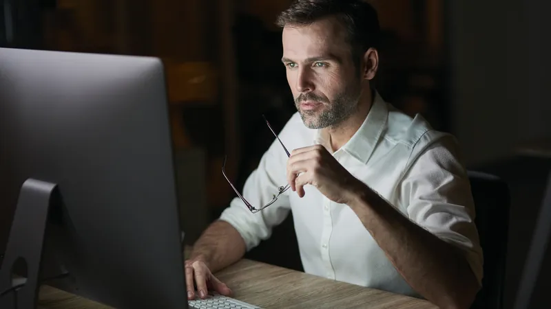 Homem concentrado trabalhando no computador à noite, segurando os óculos